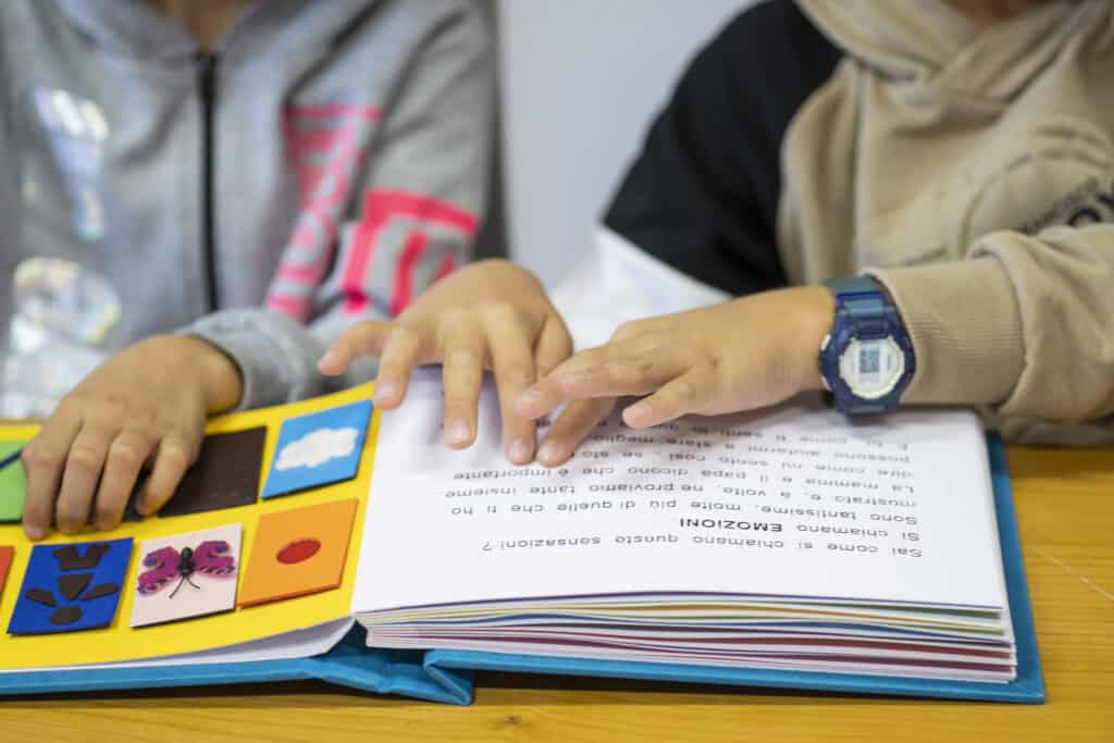 le dita di un bambino scorrono le righe di un libro illustrato tattile per leggere il braille. Accanto a lui, sullo sfondo una bambina tocca le figure dello stesso libro.