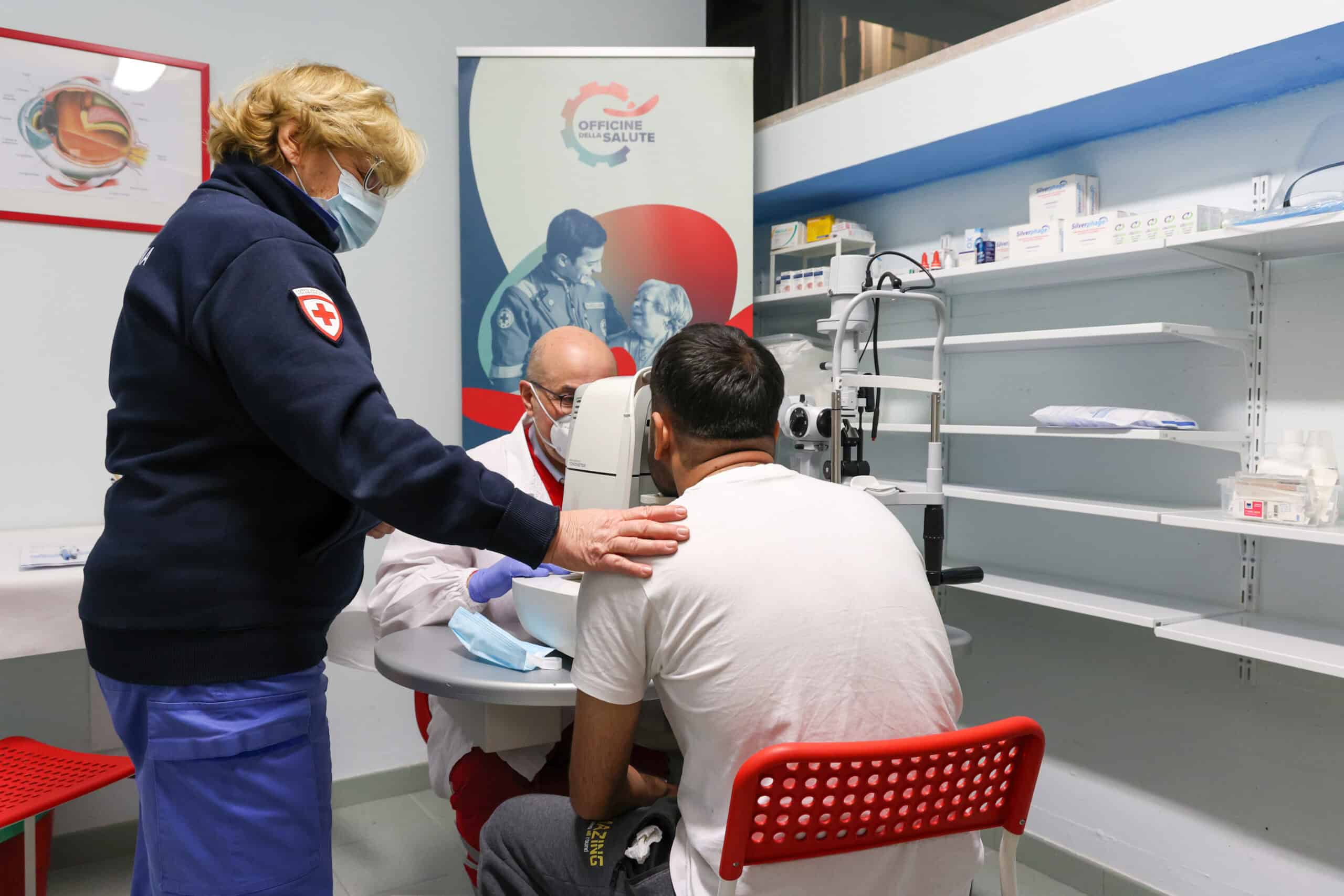 All'interno di un ambulatorio un medico effettua su un paziente il controllo della vista. Una volontaria della Croce Rossa tiene la mano sulla spalla del paziente per tranquillizzarlo.