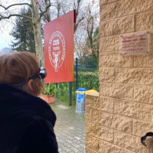 Formazione esperienziale del progetto sensoriAbile anch'io agli impianti sportivi del Cus Padova. Una donna con gli occhialini che simulano l'ipovisione cerca di leggere le parole in bordeau su una targa trasparente affissa su un muro marrone chiaro.