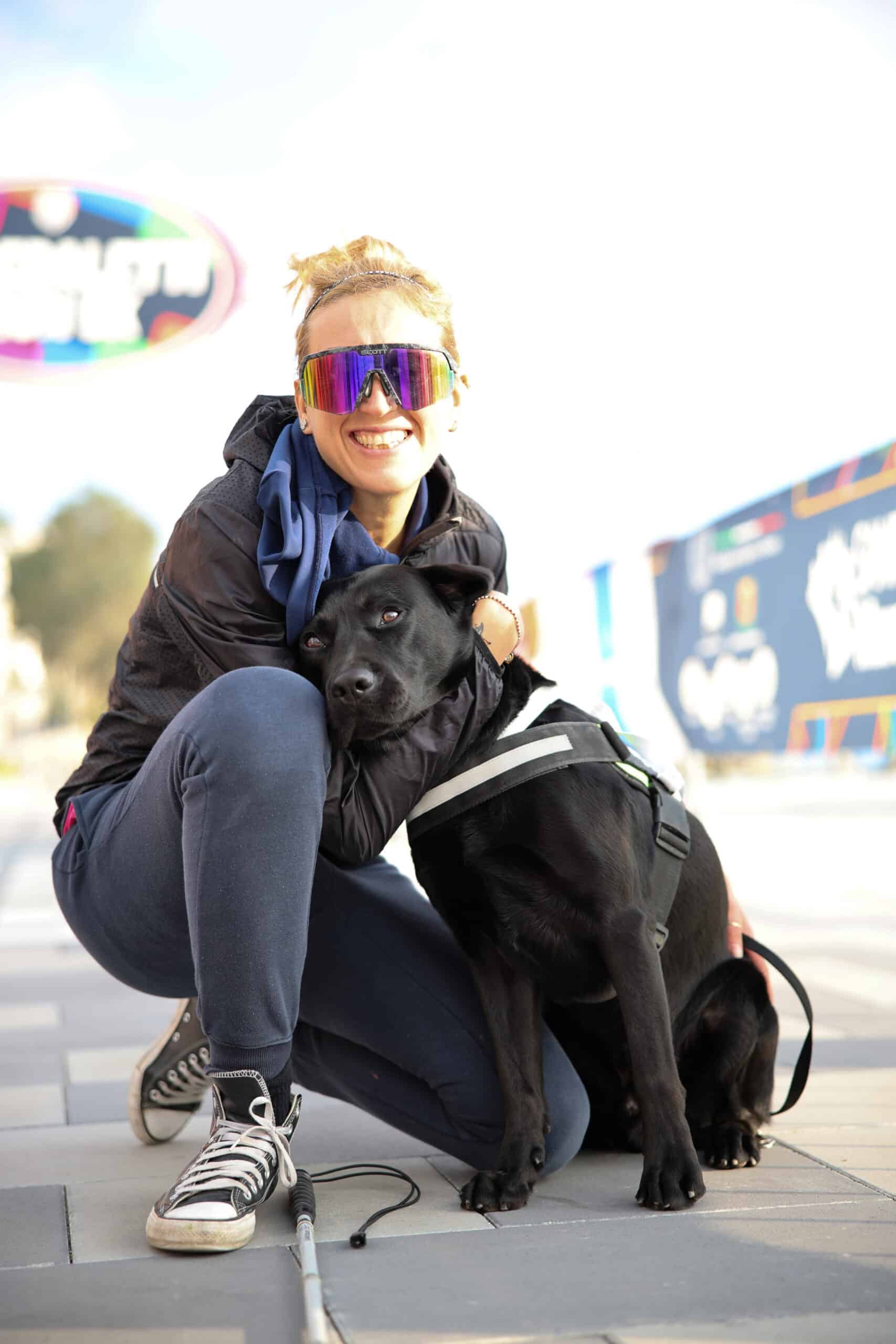 Una ragazza sorridente con occhiali scuri da ipovedente abbraccia accucciata il suo cane guida, un labrador nero.