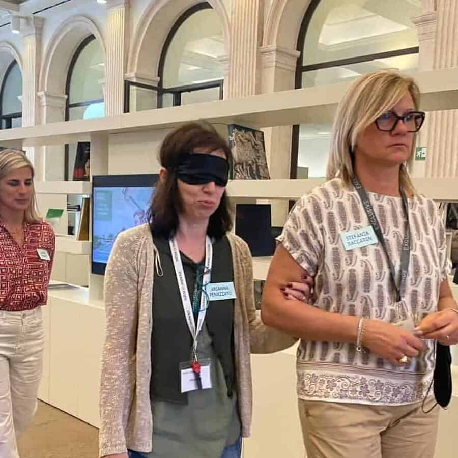 Formazione esperienziale del progetto sensoriAbile anch'io all'interno di una filiale di Intesa San Paolo. Una donna bendata cammina al braccio di una collega che la guida all'interno degli spazi della filiale. Dietro di loro c'è un'istruttrice all'orientamento e alla mobilità. 