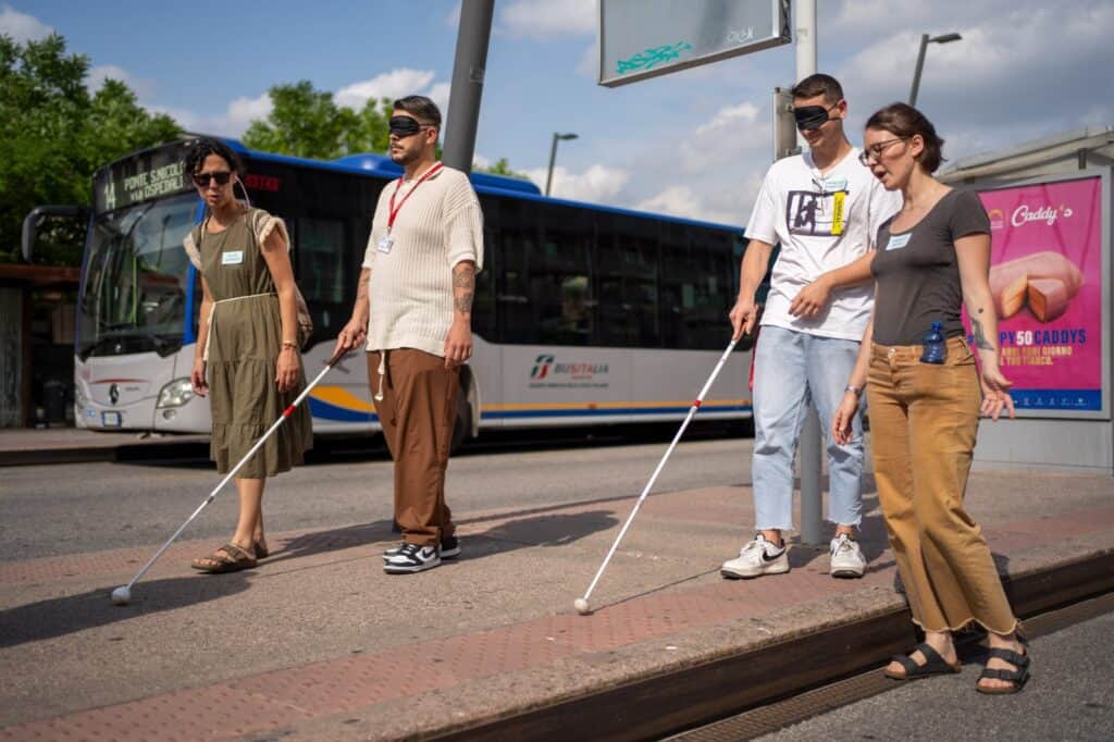 Formazione esperienziale del progetto sensoriAbile anch'io per i futuri autisti di Bus Italia Veneto.
La scena si svolge lungo una pensilina degli autobus in piazzale Stazione a Padova. Due autisti in formazione sono bendati e usano il bastone bianco cercando i logges a terra per orientarsi. Accanto a loro un'istruttrice di orientamento e mobilità e una psicologa li guidano nel percorso.