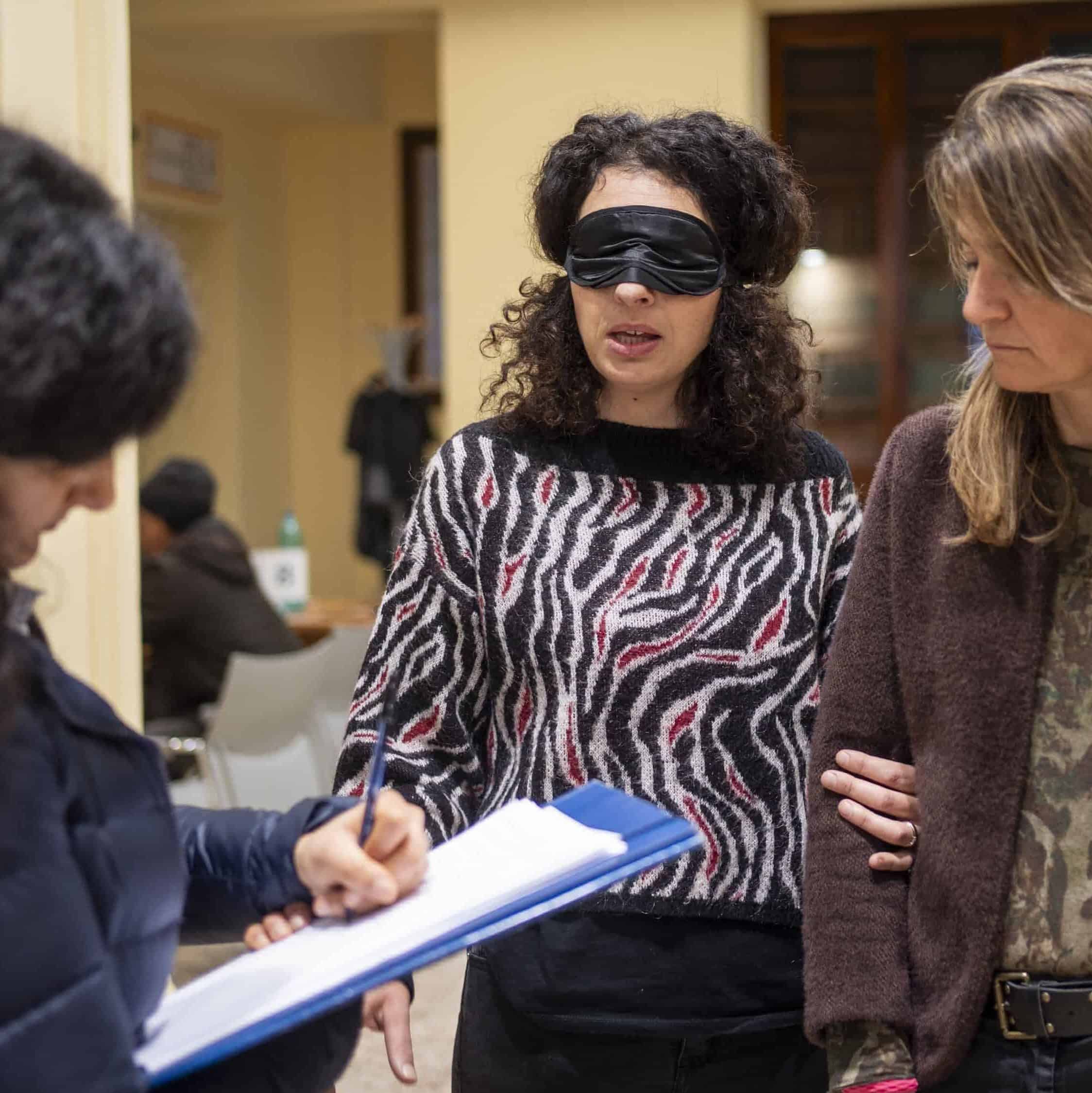 È una scena di simulazione di deprivazione visiva del progetto SensoriAbile anch'io. Siamo all'interno di un ufficio pubblico. Una ragazza bendata con una mascherina nera sta parlando, mentre tiene il braccio di una donna accanto a lei. In primo piano un'altra donna prende appunti su un foglio appoggiato a una cartellina di cartone blu.