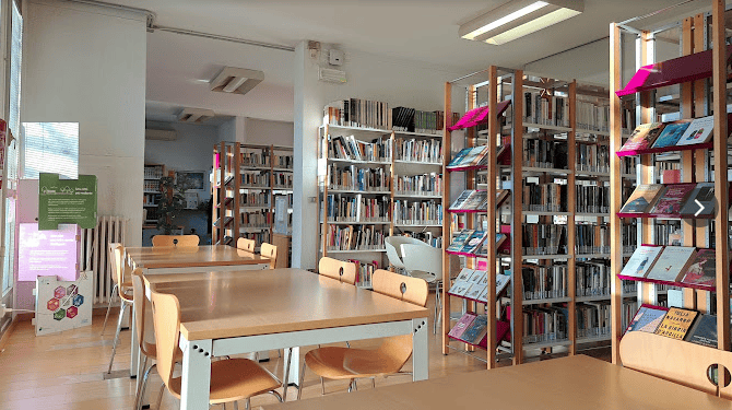 Interno della biblioteca Bassanello: tavoli e sedie in legno a sinistra dell'immagine. A destra corsie con scaffali di libri.